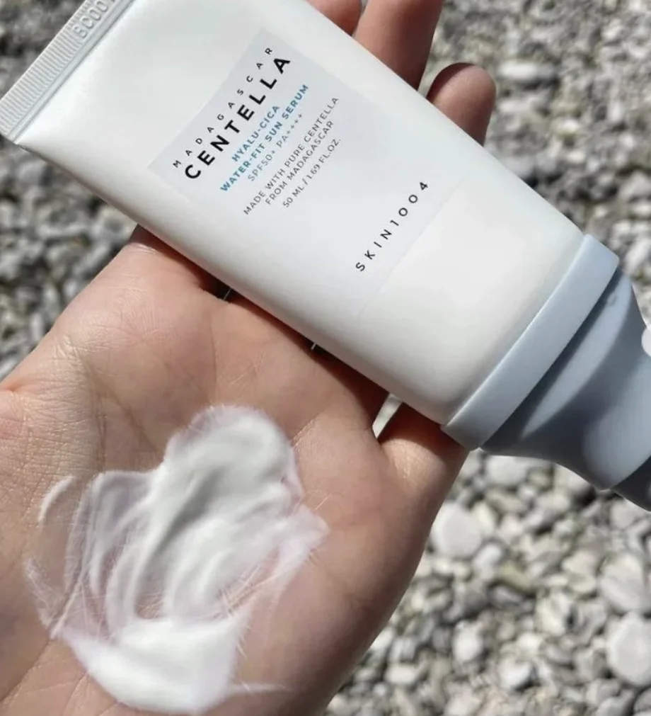Uma mão segura um tubo de protetor solar branco da marca SKIN1004 Madagascar Centella. Na palma da mesma mão, há uma amostra do produto, que possui uma textura cremosa e fluida de cor branca.