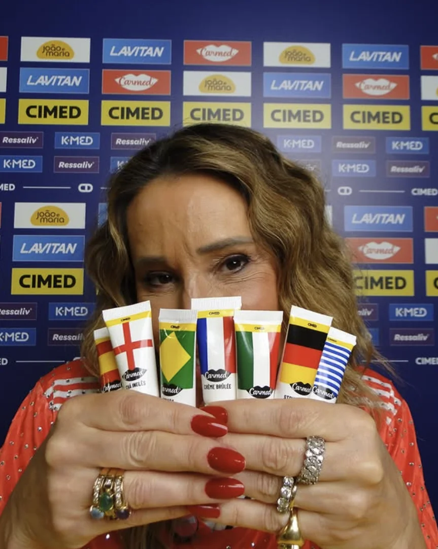 close-up de uma mulher segurando várias bisnagas de hidratante labial Carmed, cada uma com o padrão de uma bandeira nacional diferente, na frente de um painel de logos de marcas patrocinadoras. Ela usa anéis chamativos e tem unhas vermelhas.