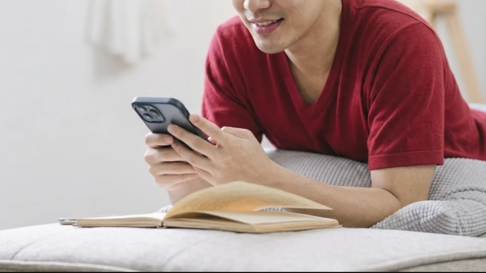Homem jovem sorrindo enquanto usa o celular, deitado de bruços com um livro físico aberto à sua frente.
