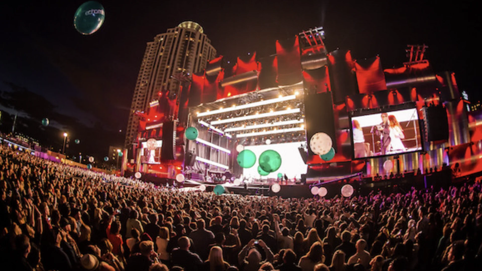 Visão panorâmica de uma grande multidão de costas, assistindo a um show em um enorme palco iluminado de vermelho e branco durante a noite. Vários balões verdes e brancos flutuam sobre o público e um grande edifício residencial se destaca ao fundo à esquerda.