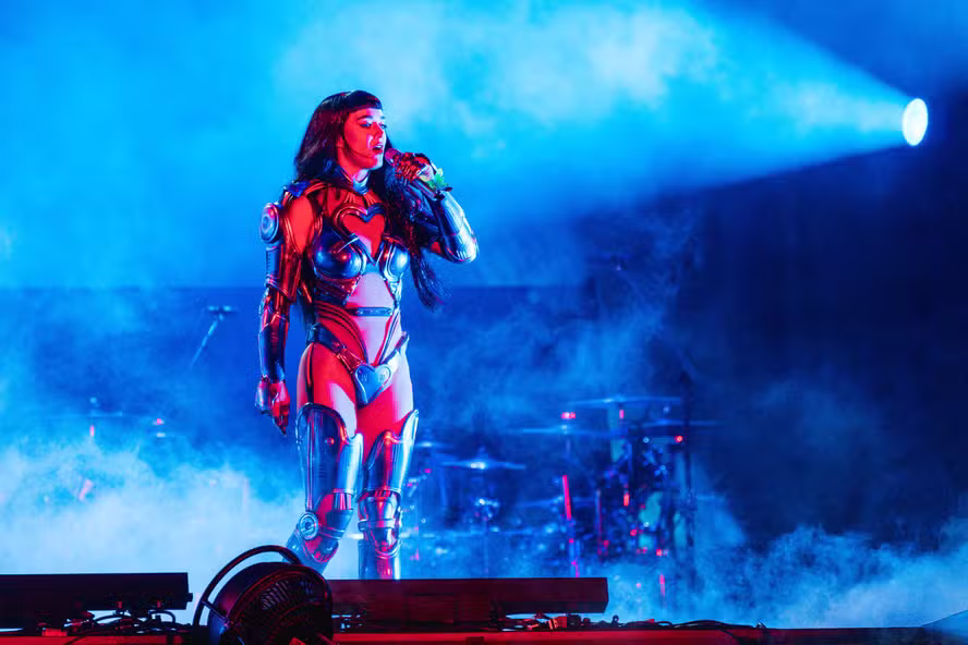 Katy Perry com cabelo curto e preto canta no palco em uma armadura futurista vermelha e prateada, cercada por luzes azuis e fumaça