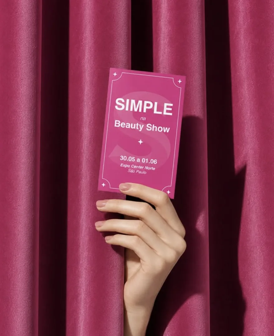 Uma mão feminina surge entre cortinas de tecido rosa-escuro segurando um cartão vertical também rosa. No cartão está escrito "SIMPLE na Beauty Show", com as datas de 30.05 a 01.06 no Expo Center Norte, São Paulo.