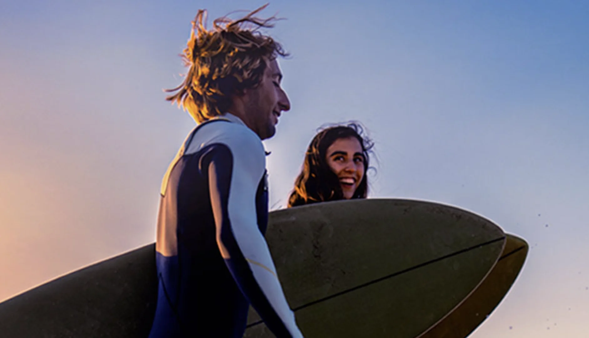 Close em perfil de um homem loiro usando roupa de borracha e uma mulher sorrindo logo atrás, ambos carregando pranchas de surfe sob o braço contra um céu claro de fim de tarde.