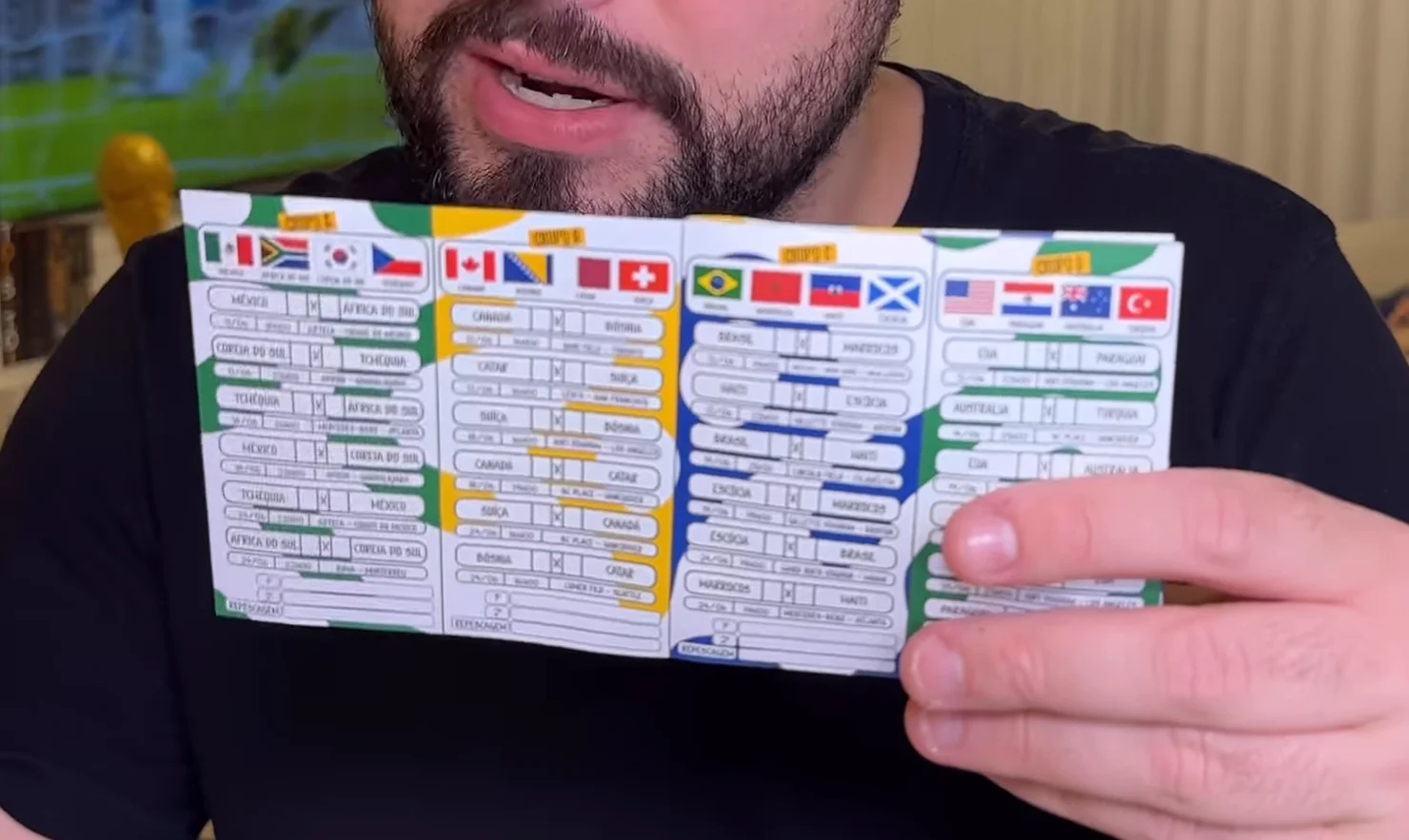Close de um folheto aberto mostrando tabelas divididas por grupos da Copa do Mundo, com bandeiras de diversos países no topo de cada coluna e espaços em branco para preencher os placares dos jogos. Um homem segura o papel com a mão direita em primeiro plano.