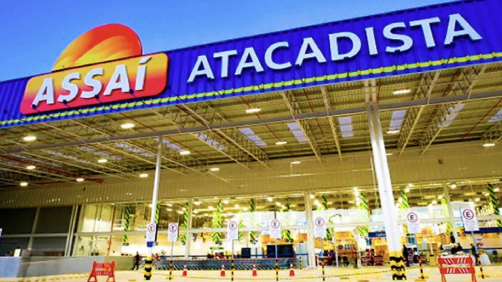 Uma vista noturna em ângulo baixo da entrada principal de uma loja Assaí Atacadista, mostrando o grande letreiro azul com "ASSaÍ ATACADISTA" e o logotipo do sol.