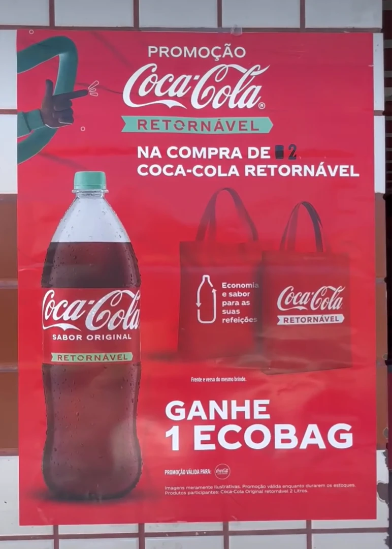 Cartaz promocional vermelho fixado em uma parede de azulejos. O texto principal diz: "Promoção Coca-Cola Retornável. Na compra de 2 Coca-Cola Retornável, ganhe 1 ecobag". À esquerda, há uma garrafa de Coca-Cola de 2 litros com tampa verde e, à direita, a ilustração das sacolas retornáveis vermelhas.