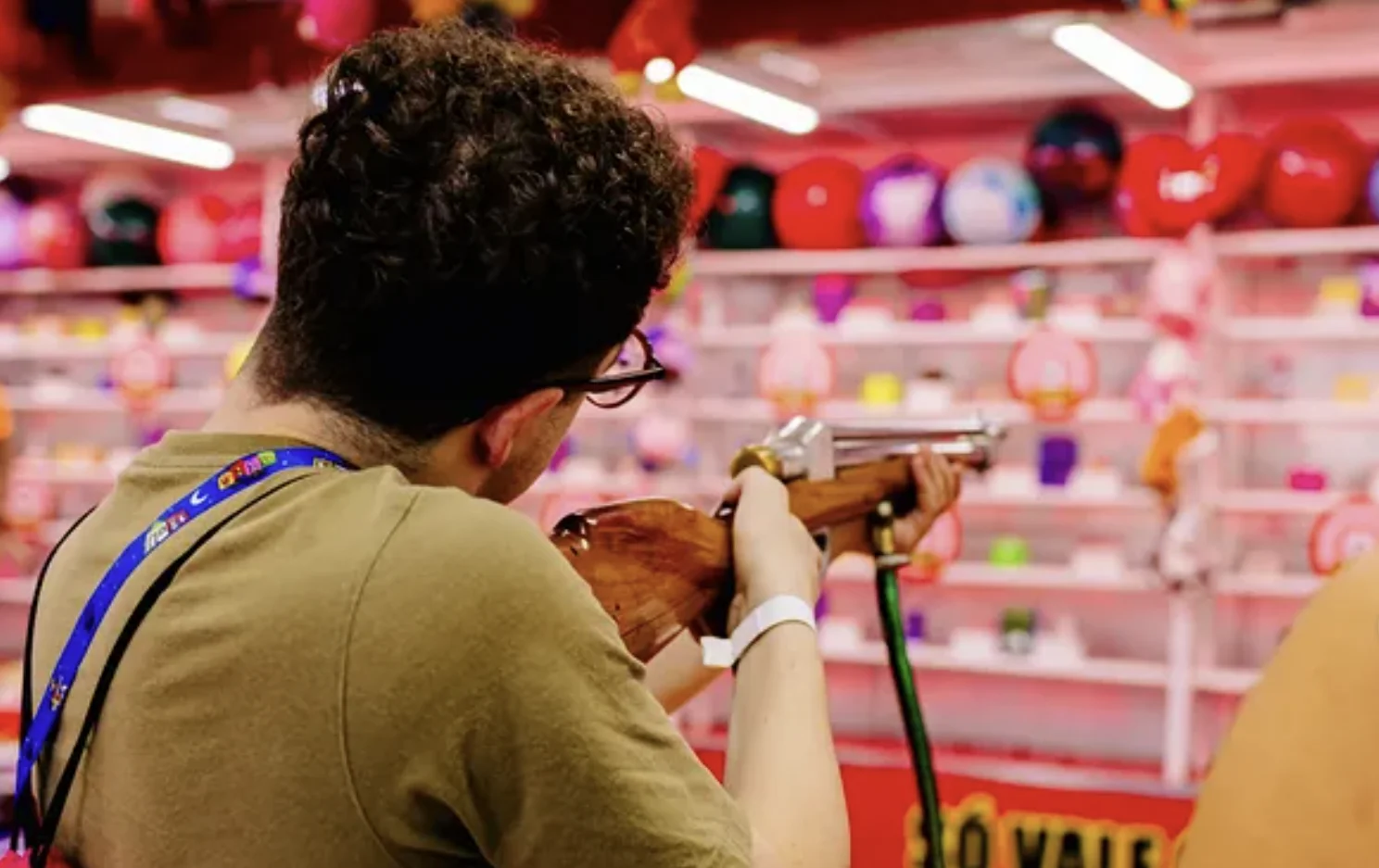 Homem de costas, usando óculos, mira com uma espingarda de brinquedo em uma barraca de tiro ao alvo colorida com prêmios e balões ao fundo.