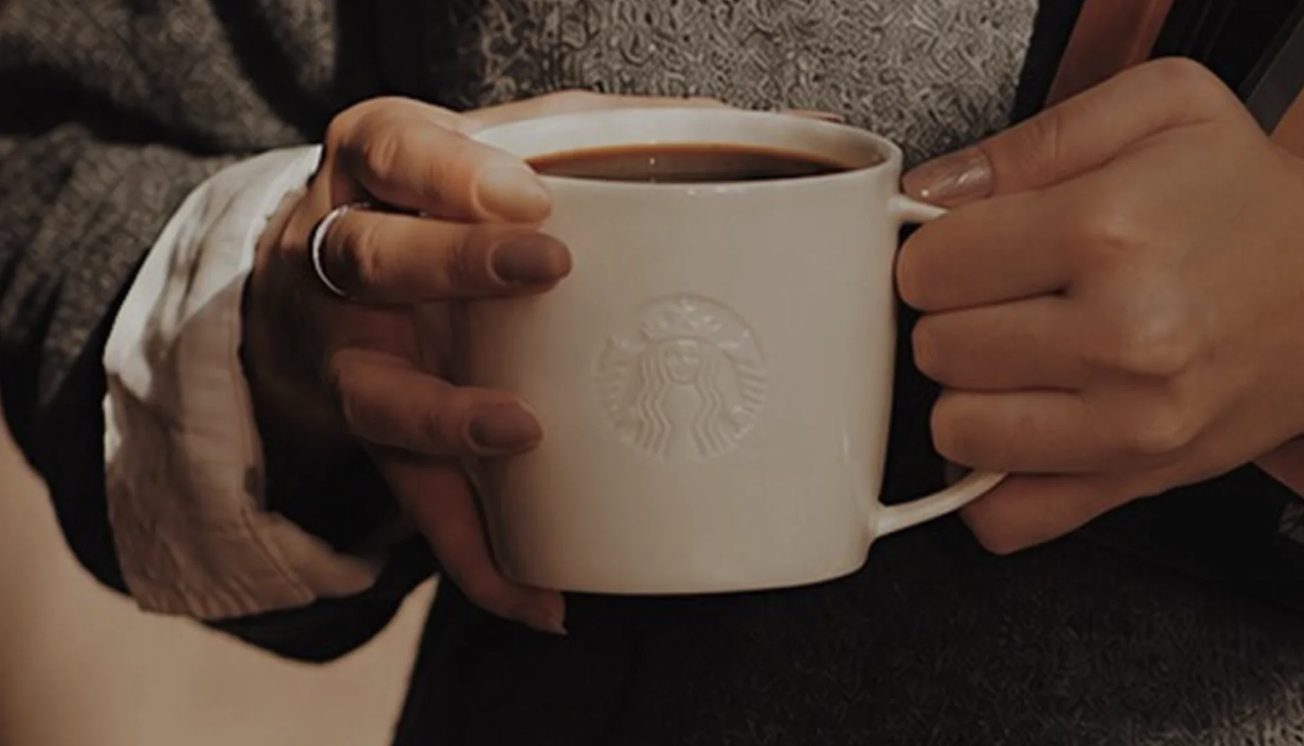 Pessoa segurando caneca branca da Starbucks com café quente em ambiente aconchegante.