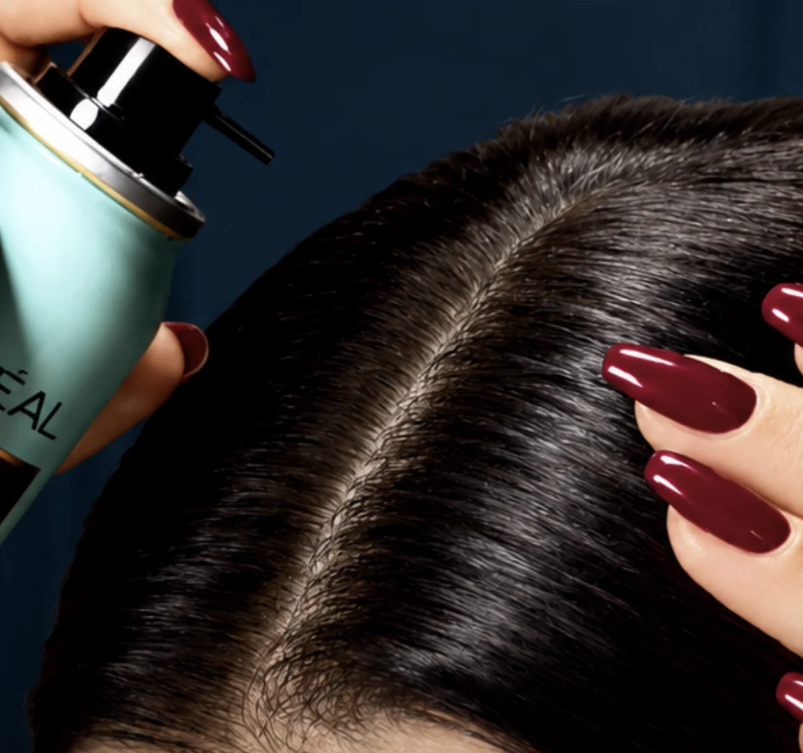 Close em uma mulher com cabelos escuros, segurando uma lata de spray L'Oréal Magic Retouch perto da raiz do cabelo, e com unhas vermelhas vibrantes.