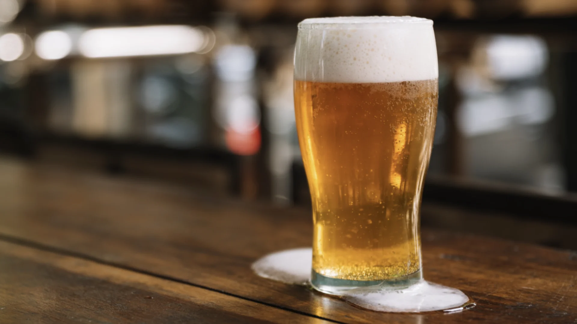 Copo de cerveja clara com espuma sobre balcão de madeira em ambiente de bar.