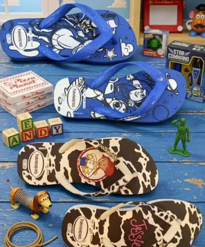 Chinelos Havaianas Toy Story 5 posicionados sobre mesa azul ao lado de brinquedos dos personagens.