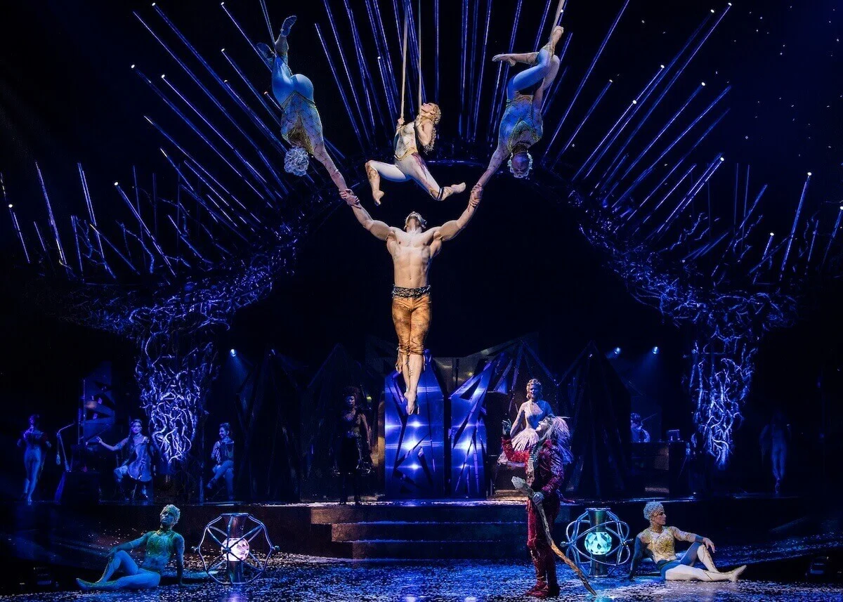 Performance de força e equilíbrio no Cirque du Soleil, onde um acrobata central segura três trapezistas suspensas no ar sob uma iluminação cênica azul e raios de luz ao fundo.