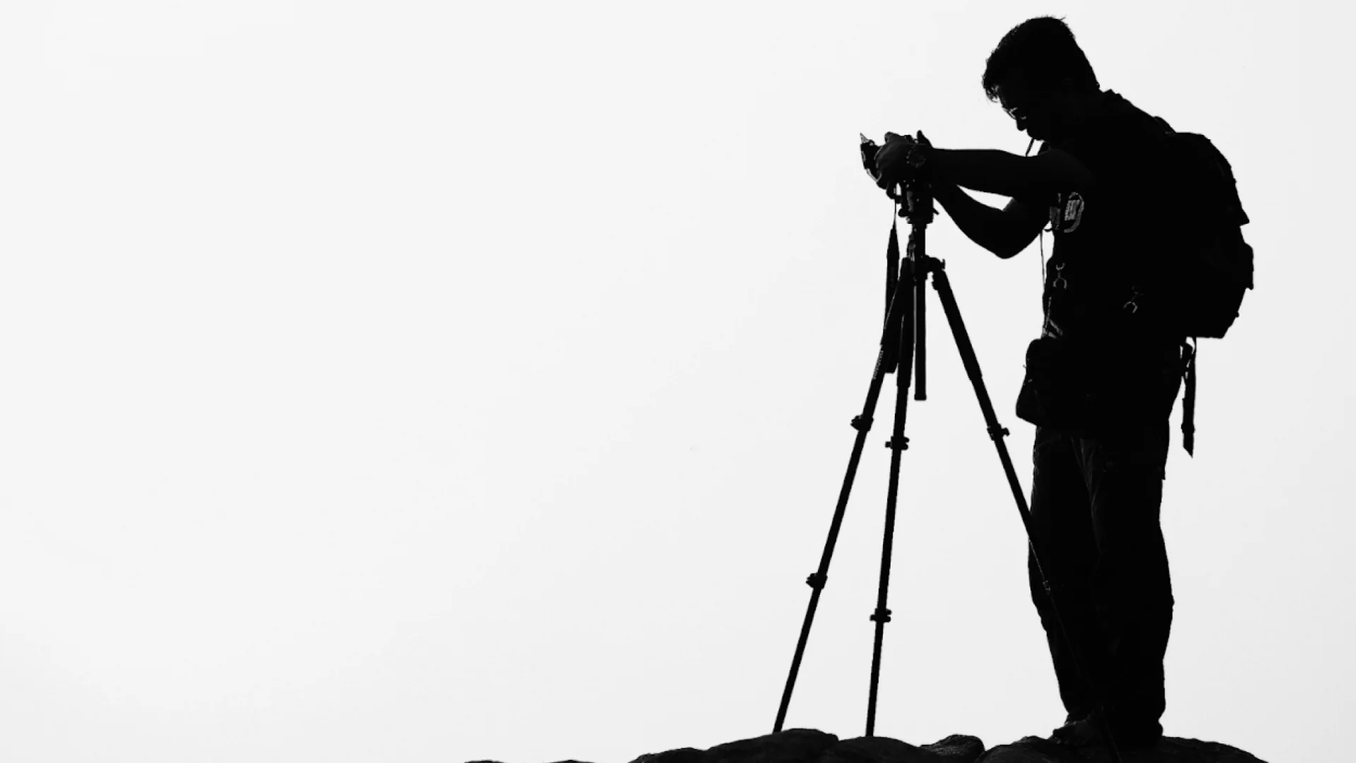 Silhueta em preto e branco de um fotógrafo de costas, ajustando sua câmera em um tripé sobre uma rocha.