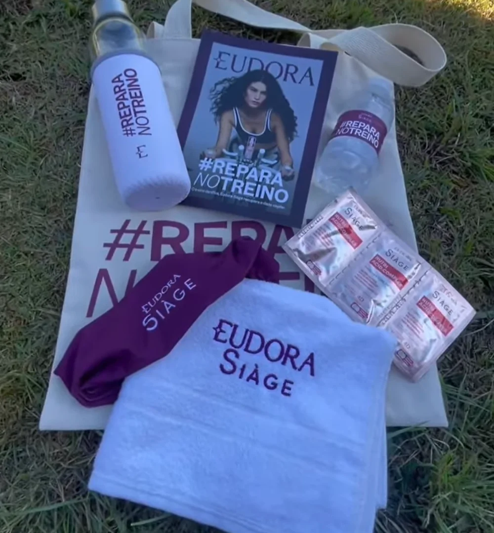 Kit da ação da Eudora Siàge com toalha, squeeze, ecobag, faixa esportiva e amostras de produtos sobre a grama.