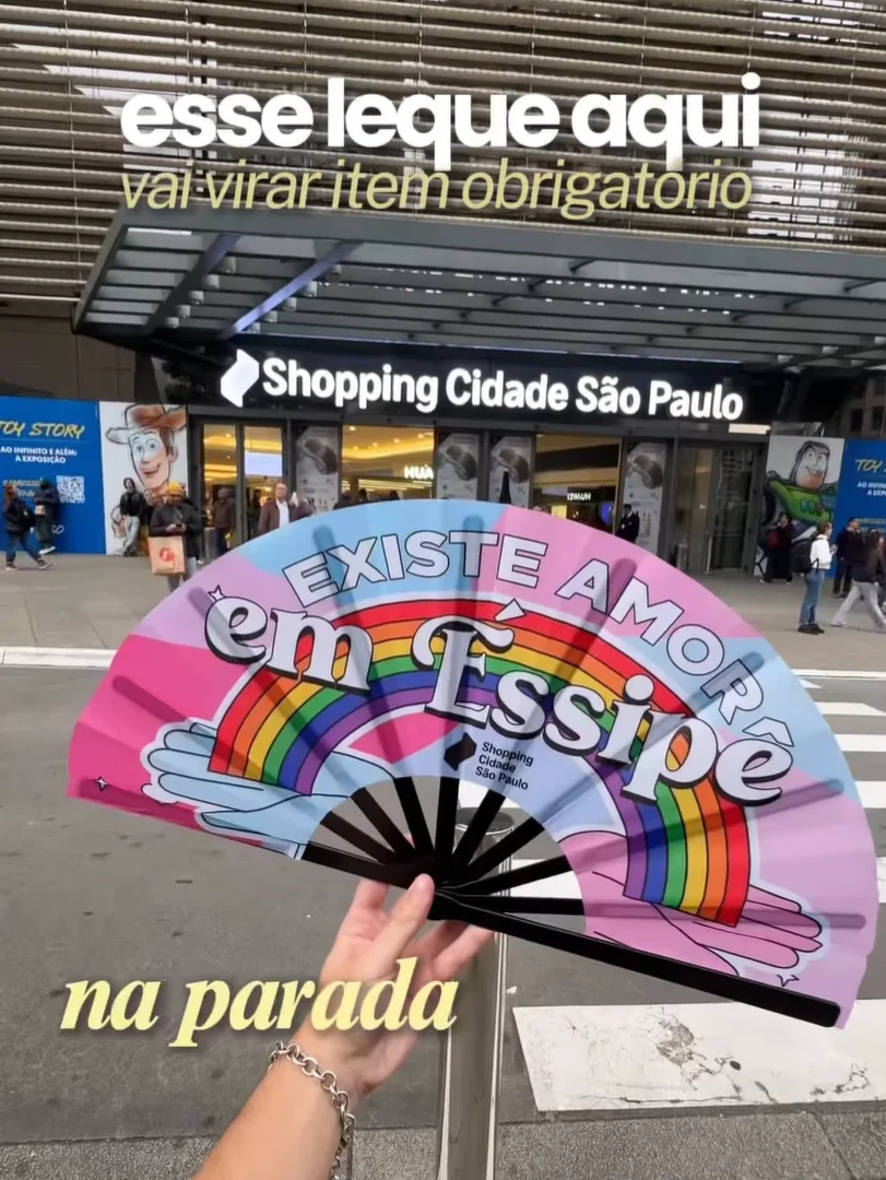 Uma mão segura um leque rosa pastel aberto com o texto preto "EXISTE AMOR em Essipe", um gráfico de arco-íris em forma de coração e o logotipo do Shopping Cidade São Paulo, posicionado em frente à entrada principal do shopping.
