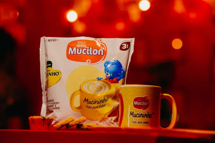 Embalagem do Mucininho ao lado de caneca personalizada em ambiente iluminado com luzes vermelhas desfocadas.