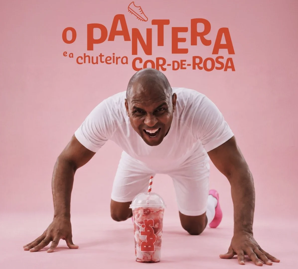 Donizete Pantera ajoelhado usando roupa branca e chuteiras rosas ao lado de milk shake do Bob’s.