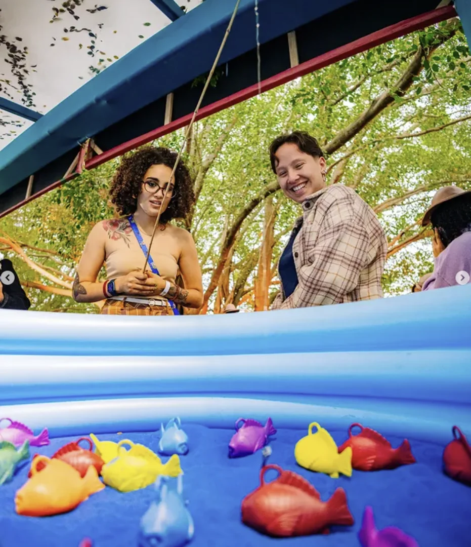 Duas pessoas sorriem perto de uma piscina inflável azul cheia de peixes de plástico coloridos na tradicional brincadeira de pescaria junina. Uma delas segura a vara de pescar.
