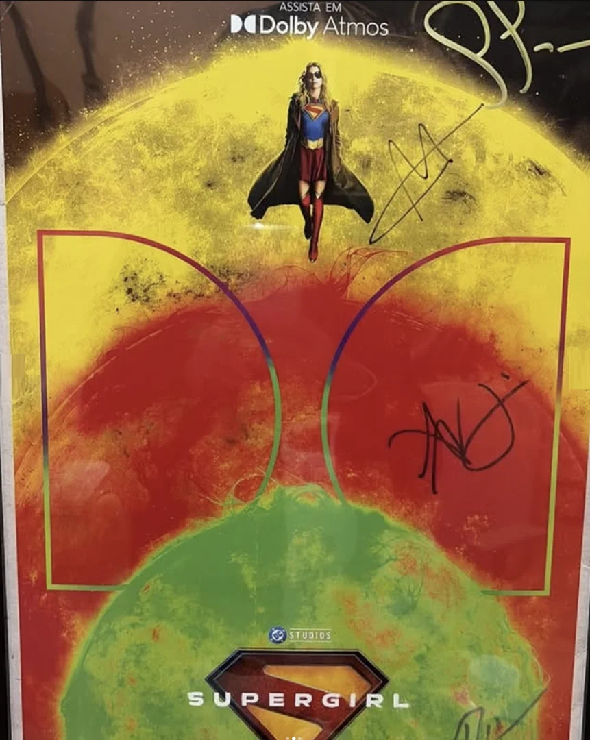 Pôster autografado de “Supergirl” com a heroína voando diante de um fundo colorido em tons amarelos, vermelhos e verdes.