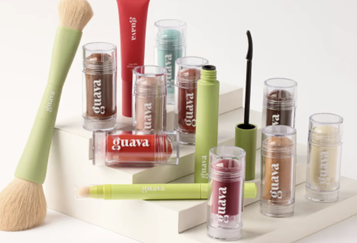 Linha de maquiagem da Guava com pincéis, gloss, máscara e sticks organizados sobre blocos claros.