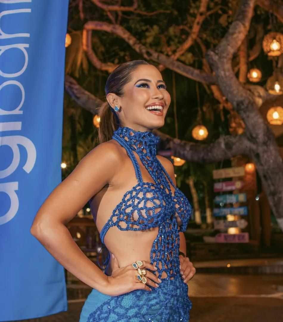 Thaynara OG sorrindo de perfil em um evento noturno, vestindo um traje azul rendado de crochê, ao lado de um banner da Agibank