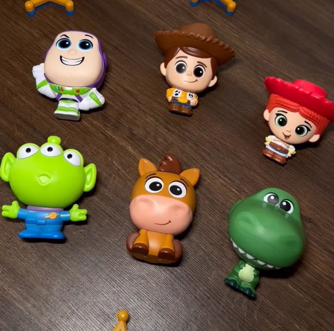 Foto tirada em ângulo superior (plongée) mostrando os seis bonecos da coleção Toy Story espalhados sobre uma mesa de madeira escura. Na fileira de cima estão Buzz Lightyear, Woody e Jessie; na de baixo, o Alien, o cavalo Bala no Alvo e o dinossauro Rex.