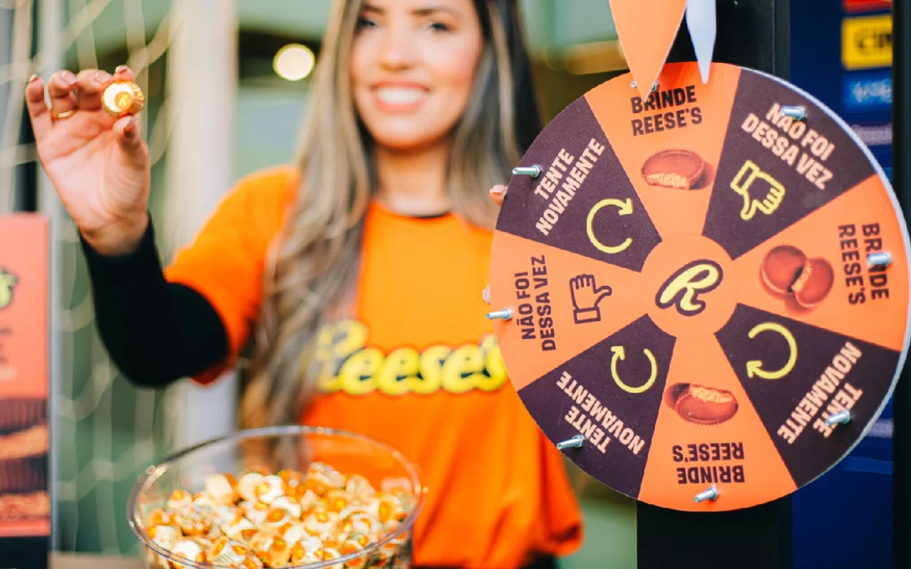 Promotora da Reese's ao lado de roleta de brindes durante ação especial do Dia Mundial do Chocolate.