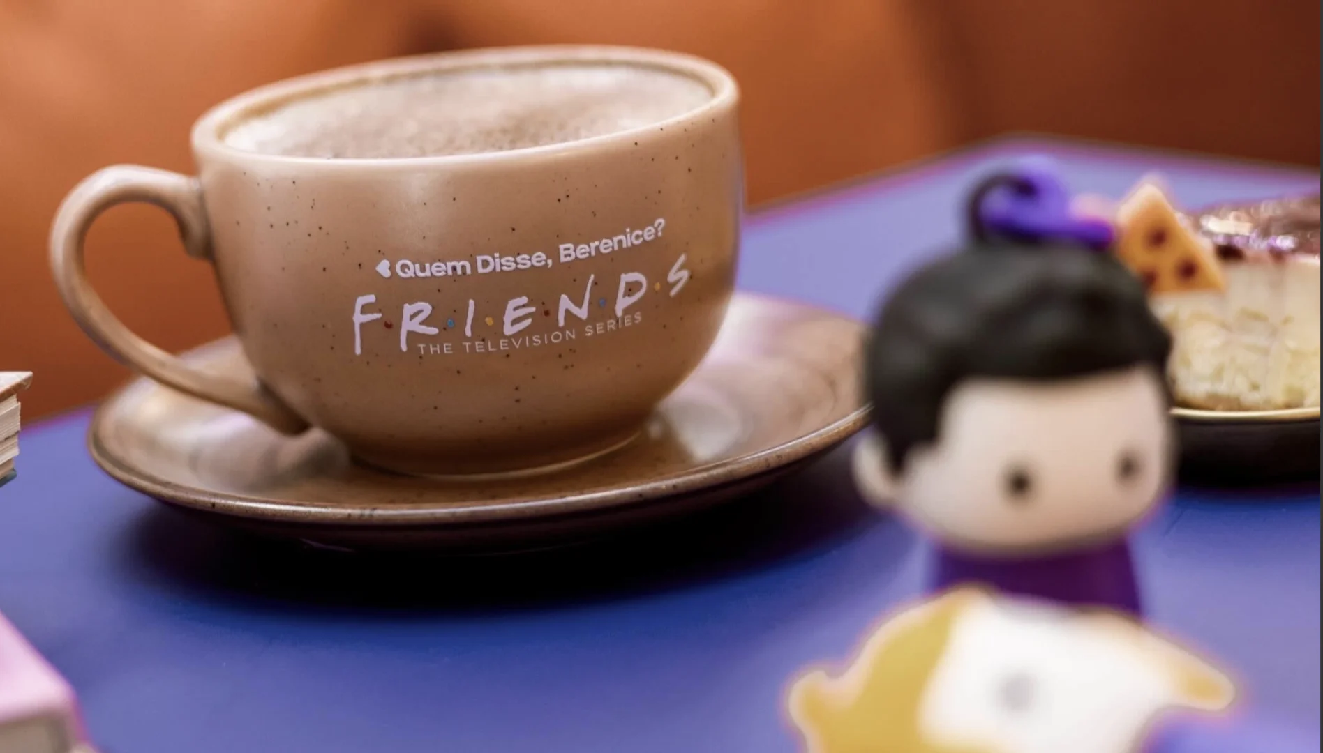 Xícara personalizada da parceria entre Quem Disse, Berenice? e Friends sobre uma mesa, com café servido e sobremesa ao fundo.