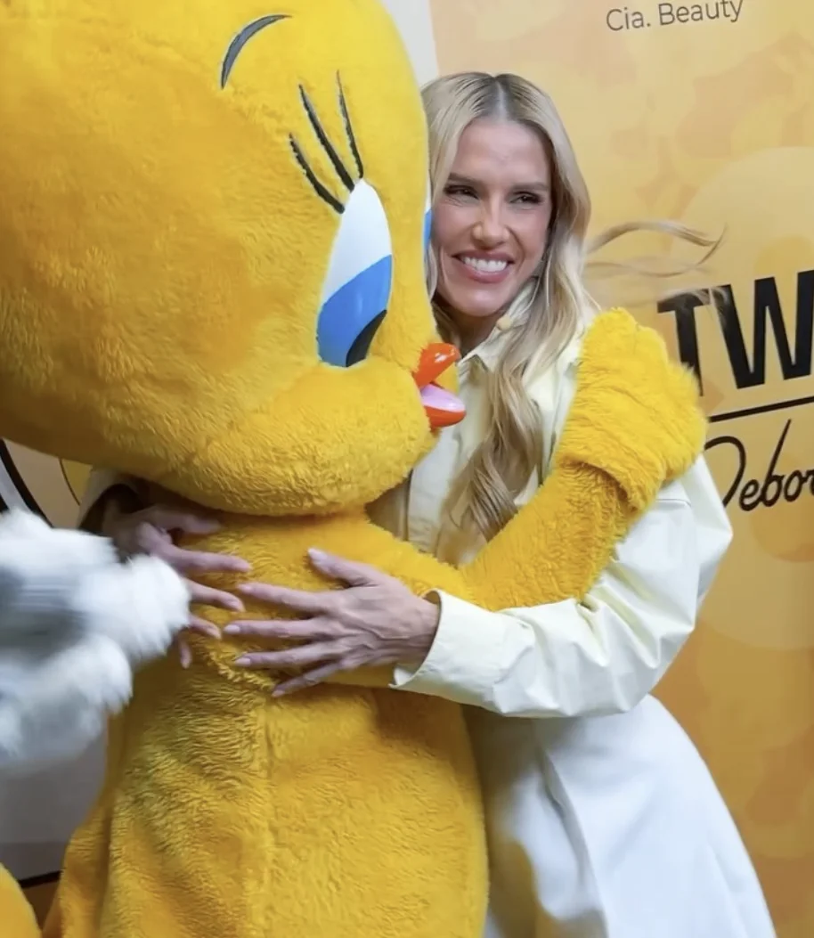 Close-up da atriz Deborah Secco, sorrindo e abraçando a personagem Piu Piu da Looney Tunes em um evento de lançamento de produto, com um fundo de marca que inclui o texto "Cia. Beauty".