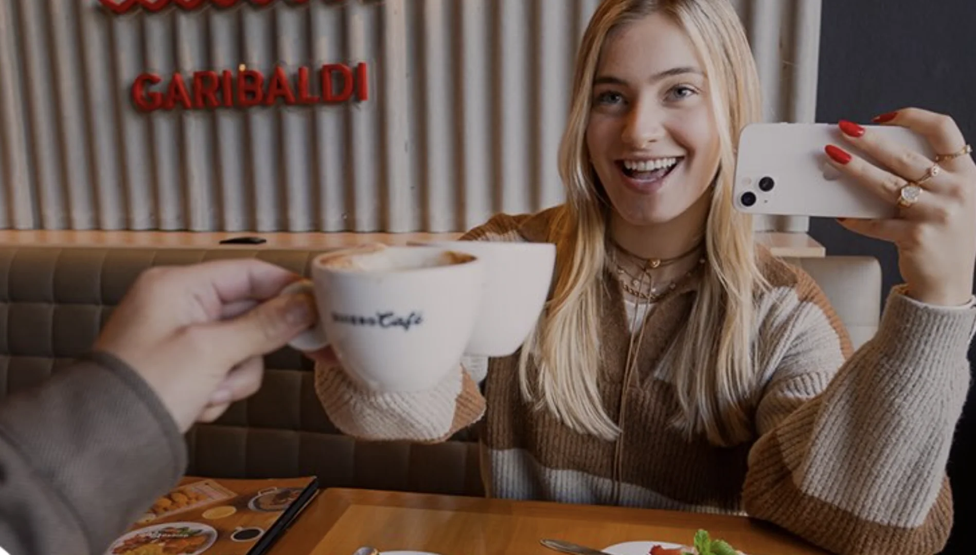 Casal brinda com xícaras de café enquanto tira uma selfie em uma cafeteria.