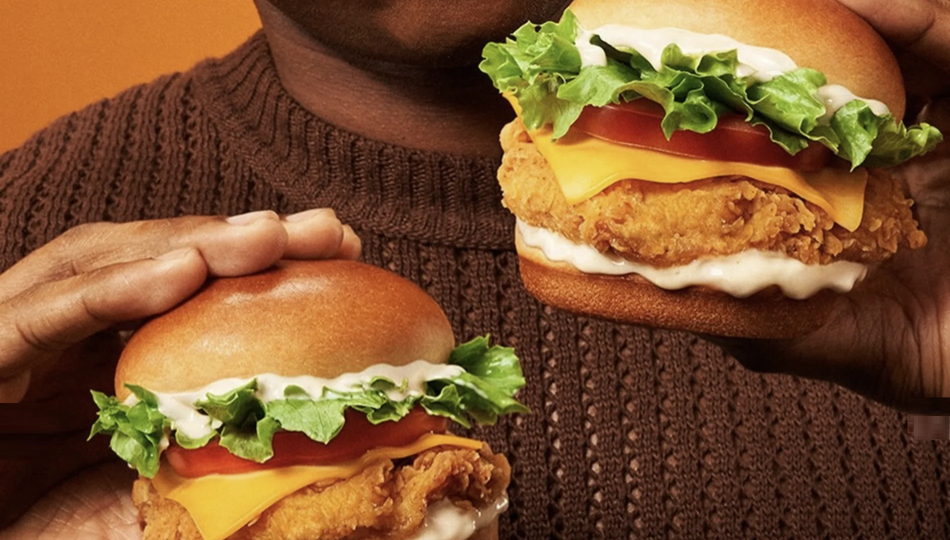 Pessoa segura dois sanduíches de frango do Popeyes com queijo, alface, tomate e molho, destacando os ingredientes da nova linha Popeyes Minis.