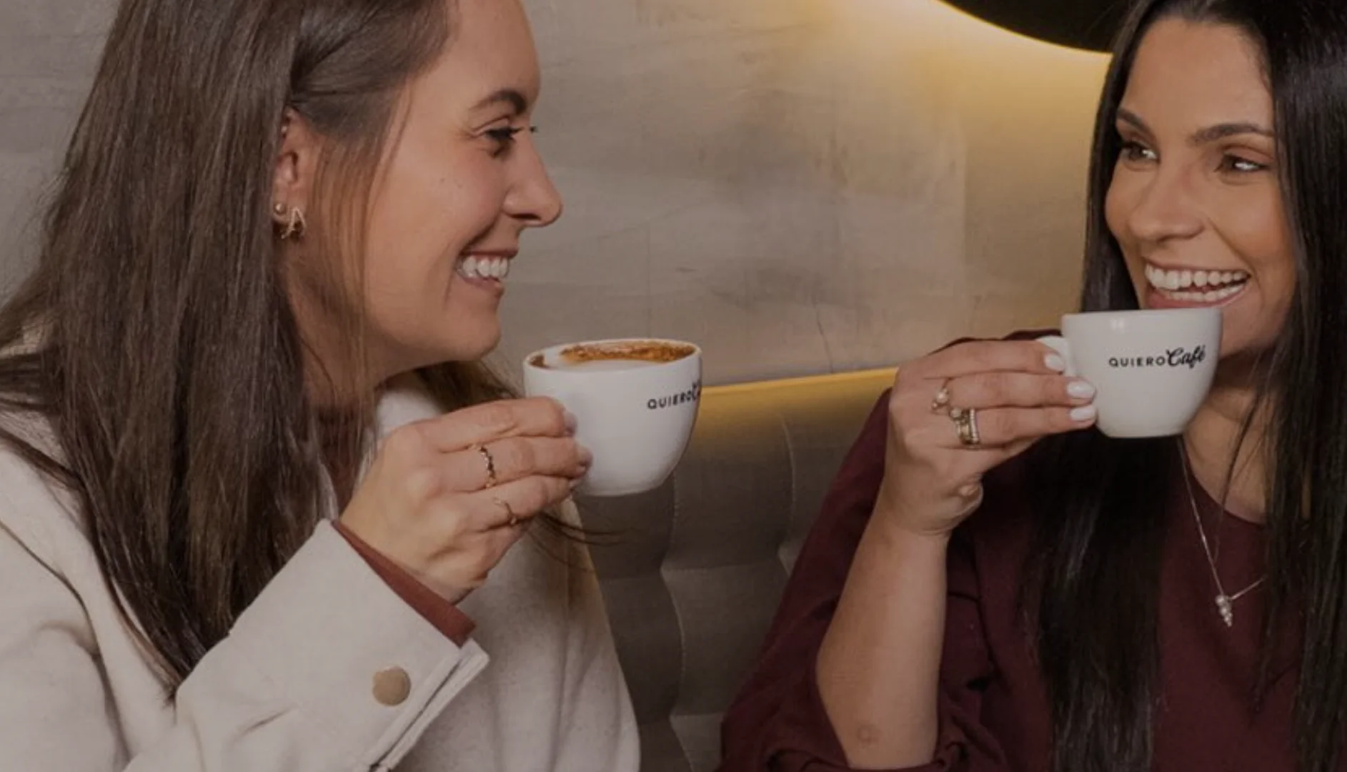 Duas amigas sorriem enquanto tomam café em uma cafeteria.