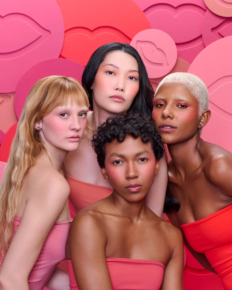 Quatro modelos com diferentes tons de pele usam maquiagem Boca Rosa diante de fundo rosa com ilustrações de lábios.