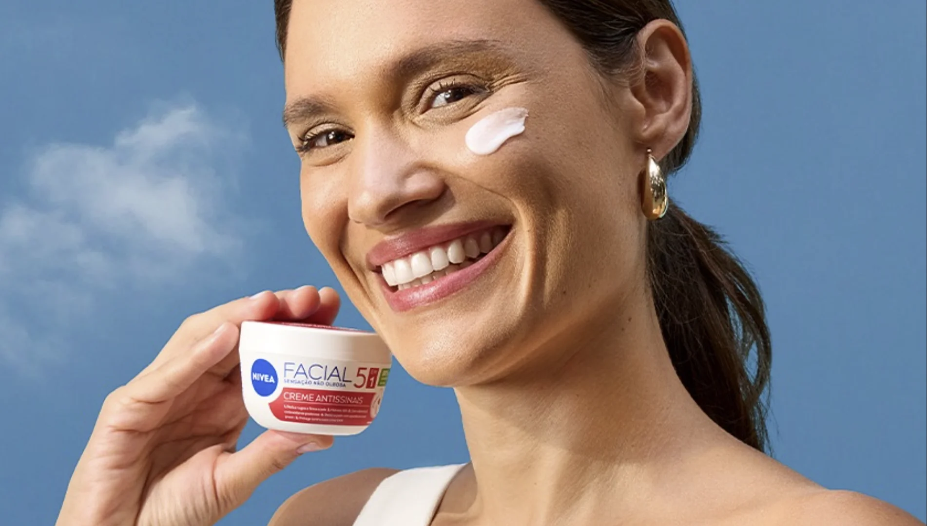 Mulher sorrindo segura um creme facial Nivea enquanto aplica o produto no rosto durante campanha da marca.