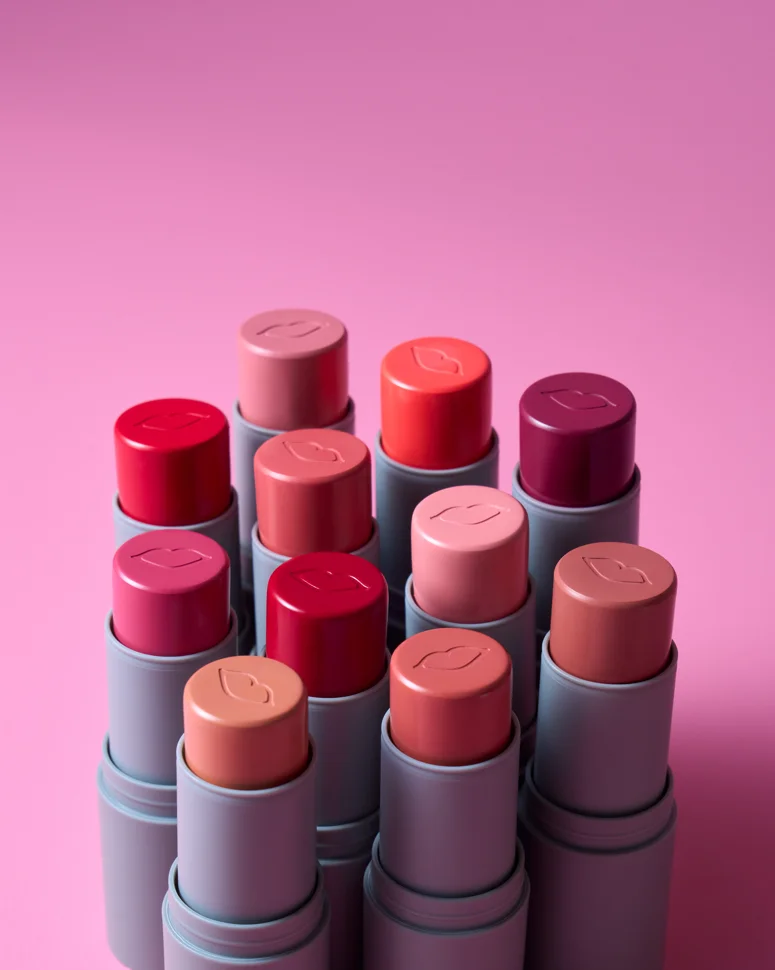 Bastões Stick Cor da Boca Rosa em diversas tonalidades de blush e maquiagem cremosa sobre fundo rosa.