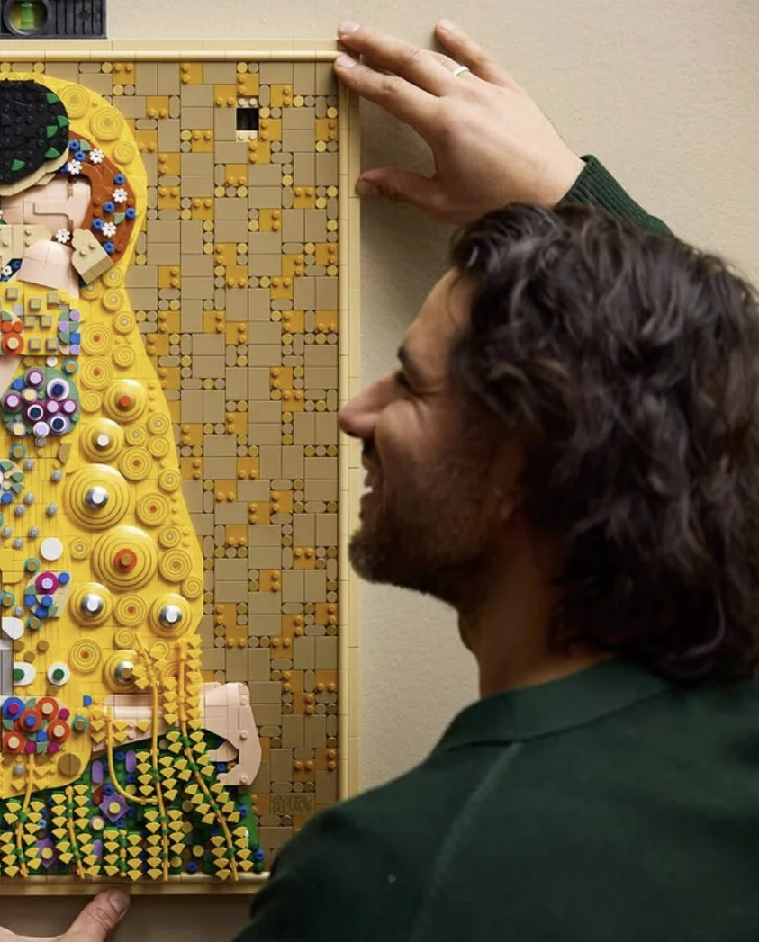 Pessoa instalando na parede o quadro LEGO Art inspirado em "O Beijo", de Gustav Klimt, após a montagem.
