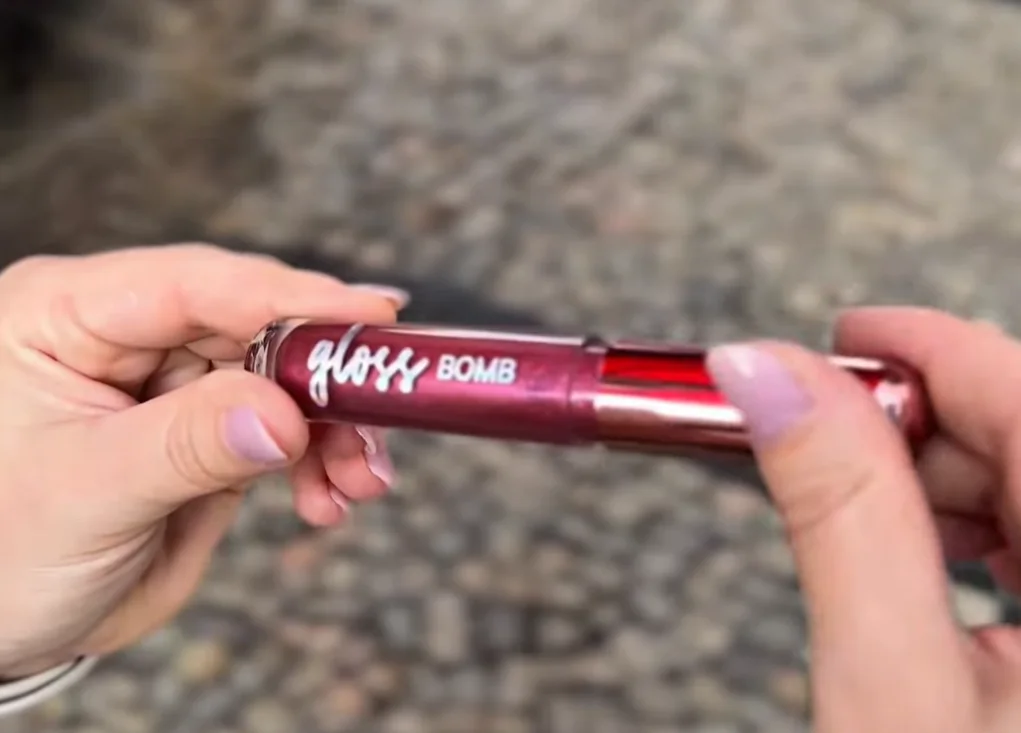 Pessoa segurando gloss labial Gloss Bomb DaBelle com fundo desfocado.