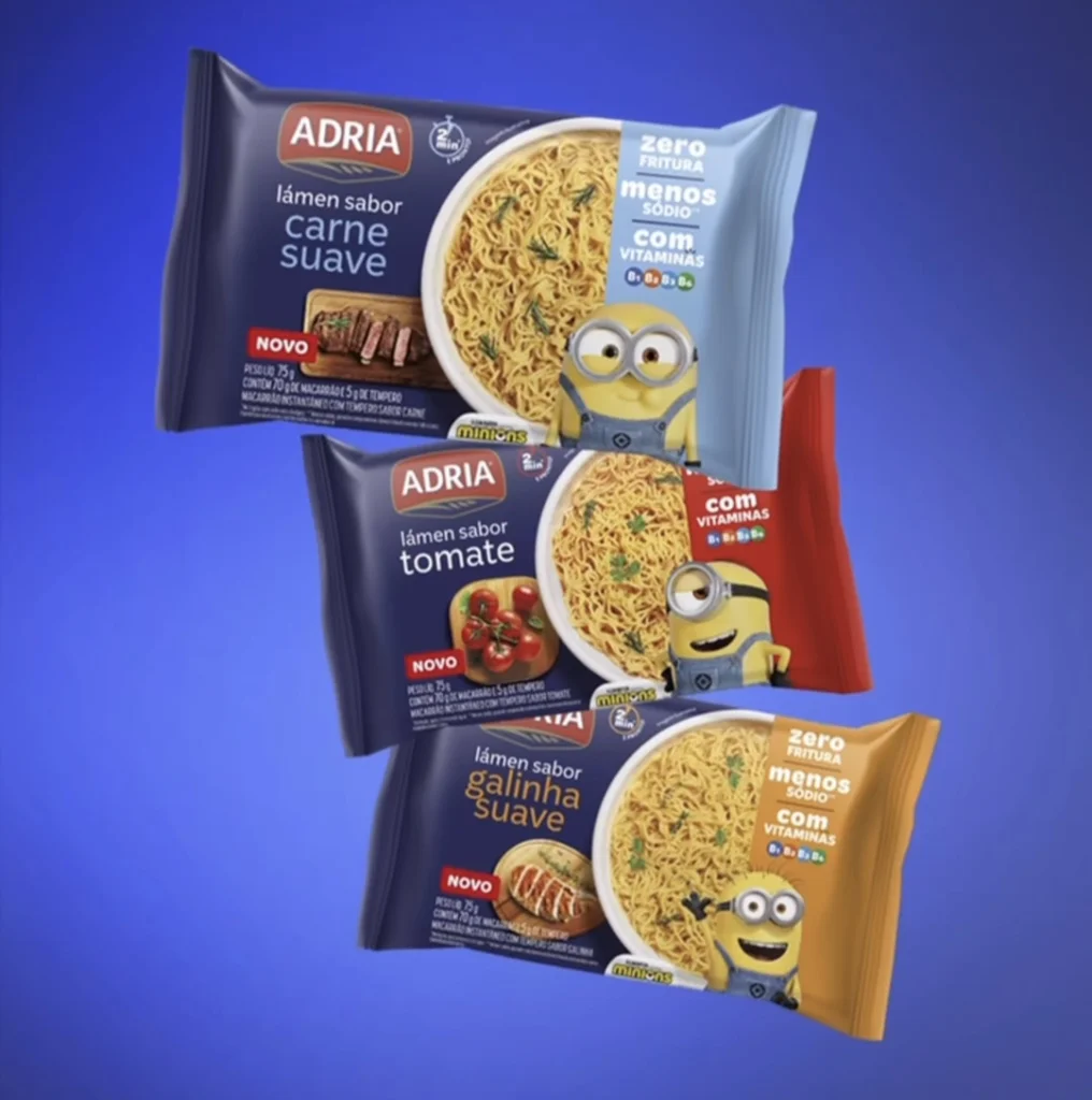 Três embalagens de macarrão instantâneo Adria com ilustrações dos Minions nas versões carne suave, tomate e galinha suave sobre fundo azul.