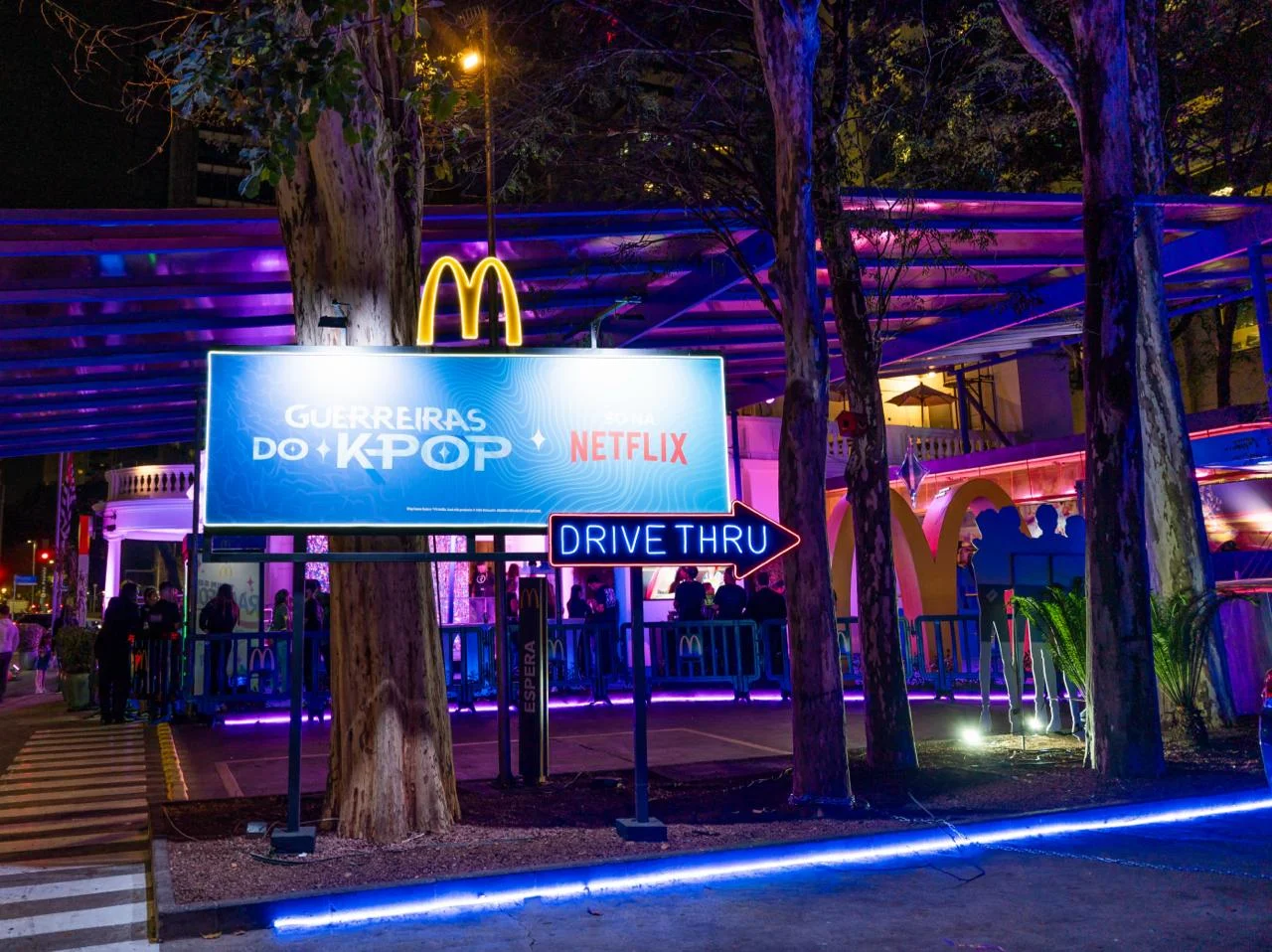 Área externa do drive-thru do Méqui 1000 com painel de Guerreiras do K-Pop e Netflix.