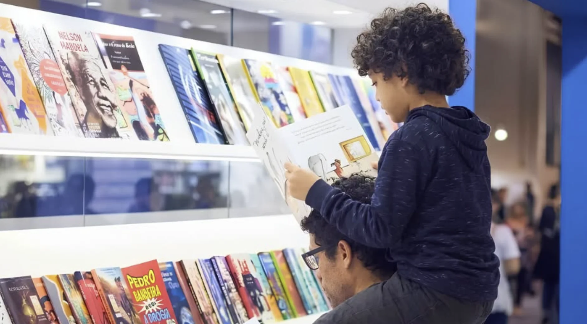 Criança lê um livro sentada nos ombros de um adulto em frente a uma estante repleta de livros durante a Bienal do Livro de São Paulo.
