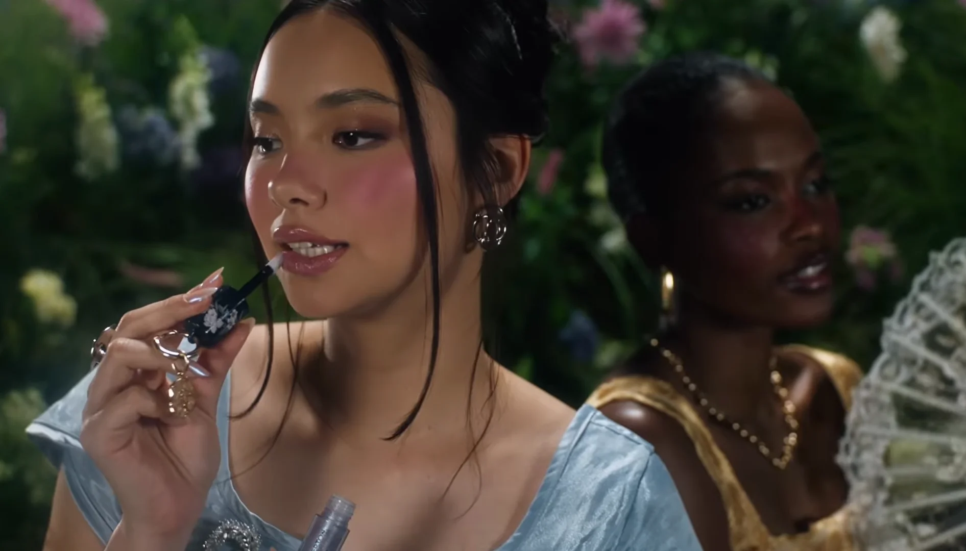 Modelo aplica gloss da coleção Bridgerton x Bruna Tavares em cenário com flores e figurino inspirado na série.