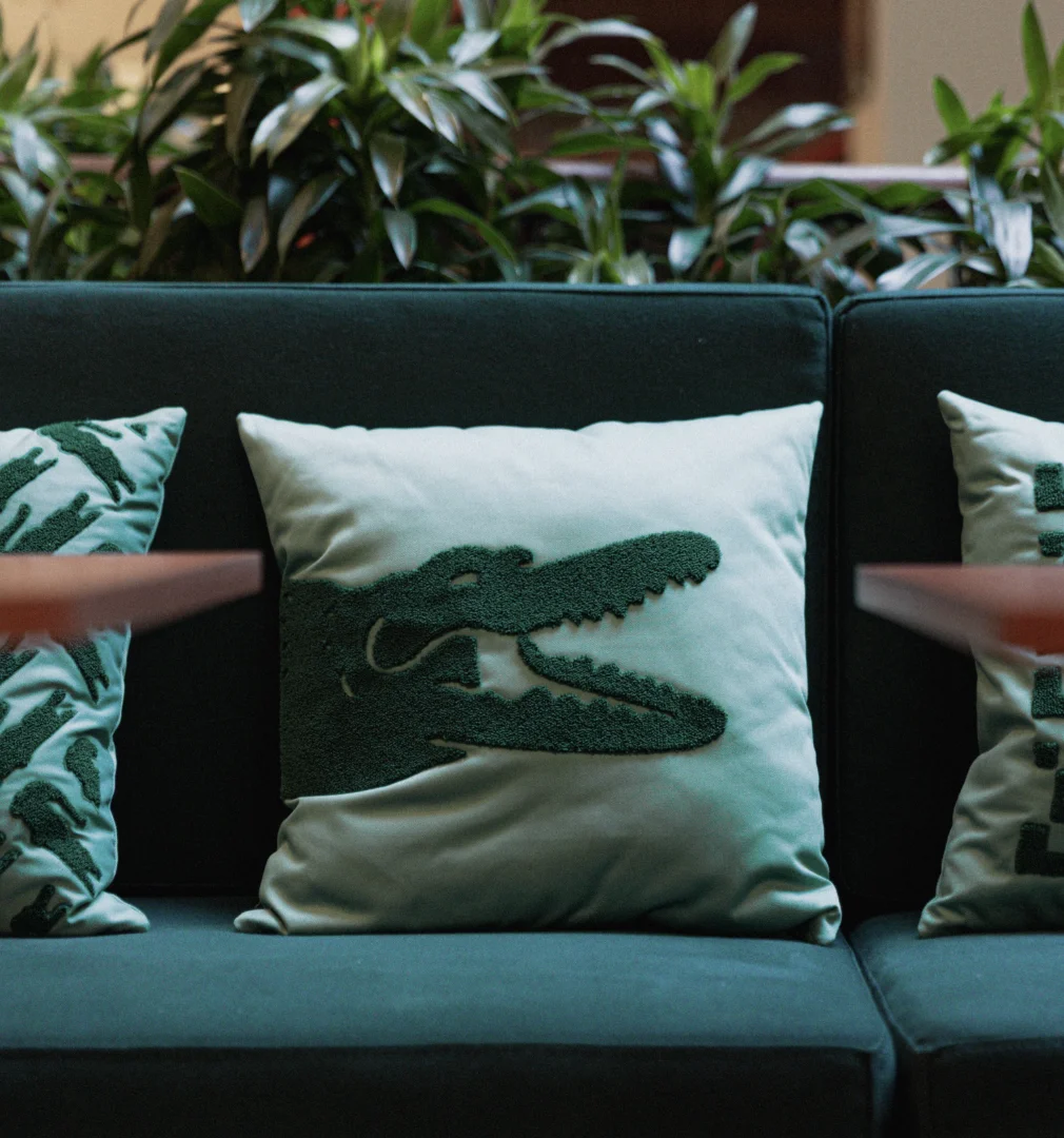 Almofada verde com bordado do crocodilo da Lacoste em sofá de ambiente interno.