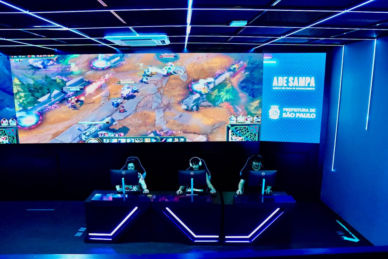 Sala gamer do Hub Sampa Games com jogadores em computadores diante de um grande telão exibindo uma partida de game, em espaço iluminado por luzes de LED na cidade de São Paulo.
