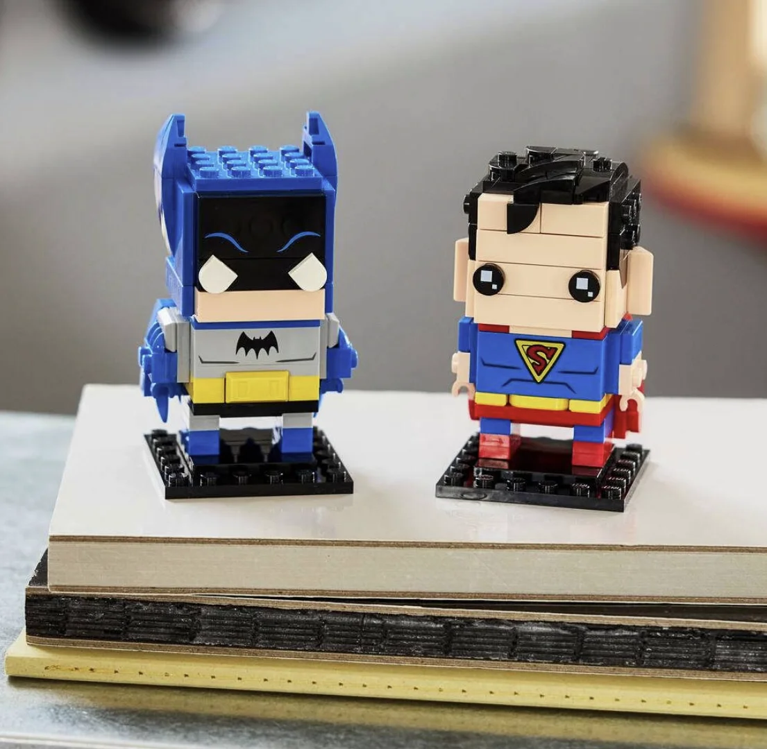 Bonecos LEGO BrickHeadz de Batman e Superman expostos sobre uma pilha de livros.