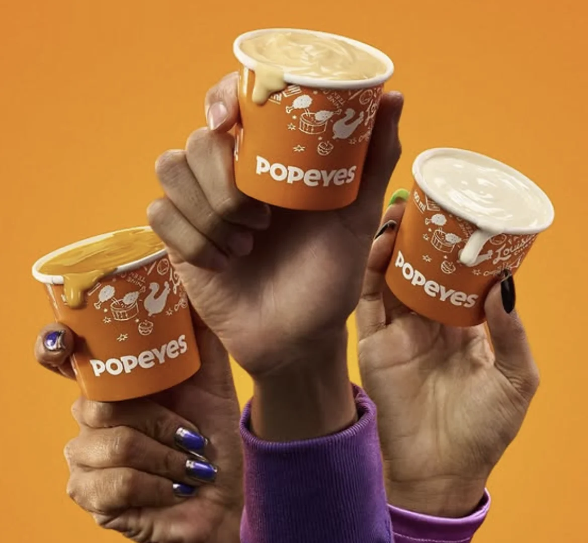 Mãos seguram três molhos de queijo do Popeyes.