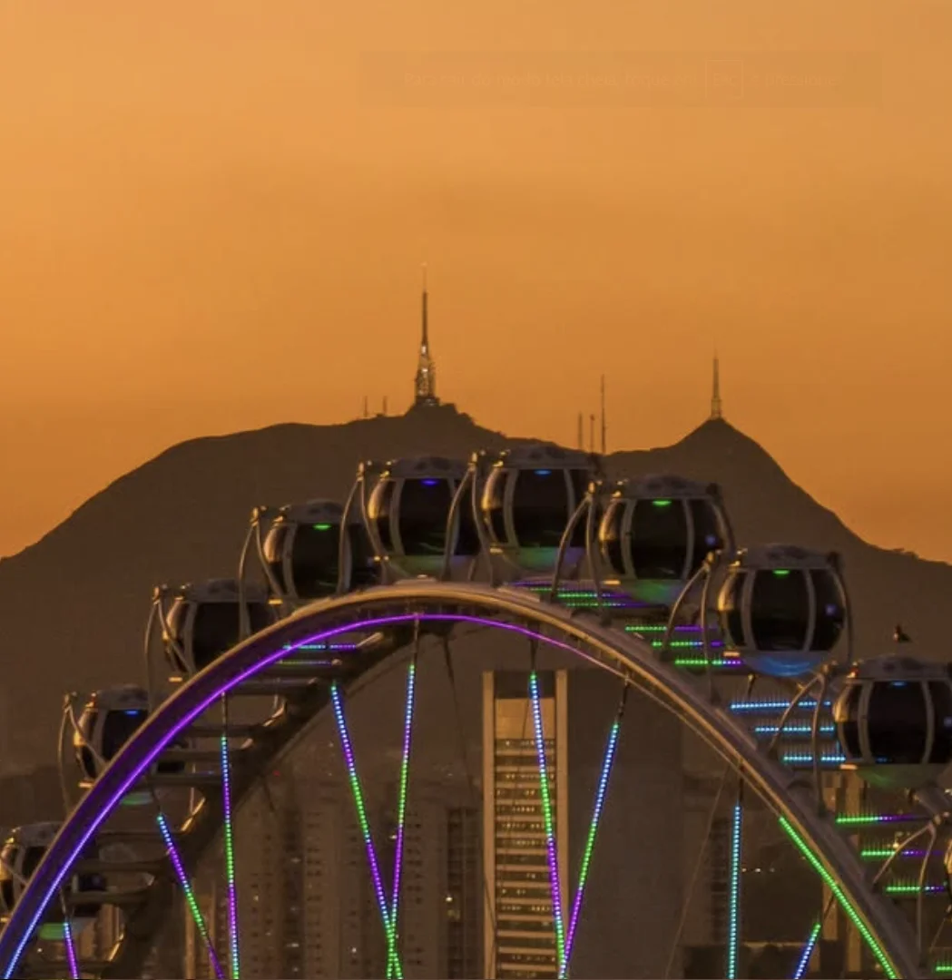 Cabines da Roda Rico iluminadas diante do céu alaranjado, com a cidade ao fundo.