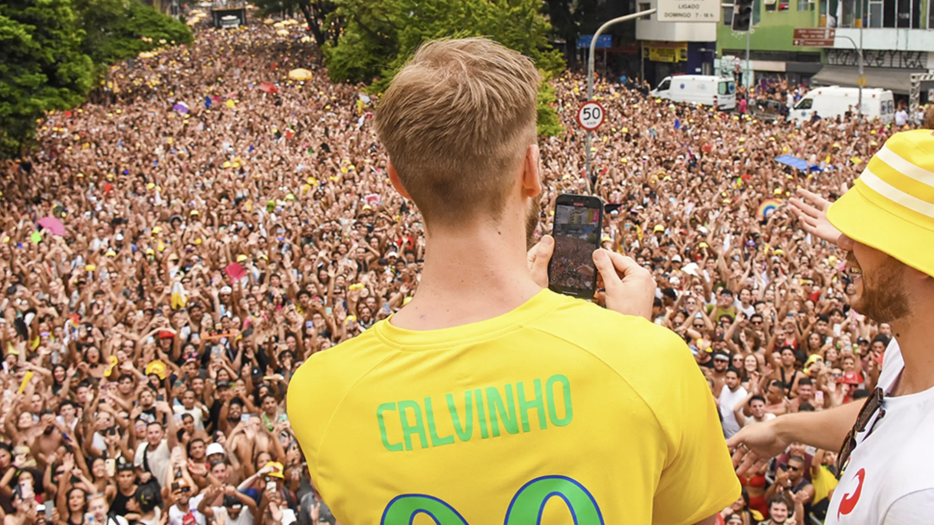 Calvin Harris vestindo uma camisa de futebol amarela com o nome "CALVINHO" nas costas está de costas para a câmera, tirando uma foto de uma multidão em uma rua em um dia ensolarado.