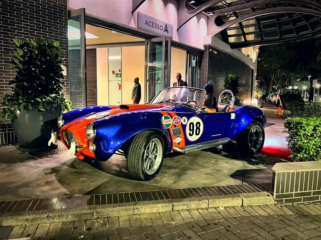 Carro clássico de corrida azul e laranja, com número 98, estacionado em frente a um edifício.