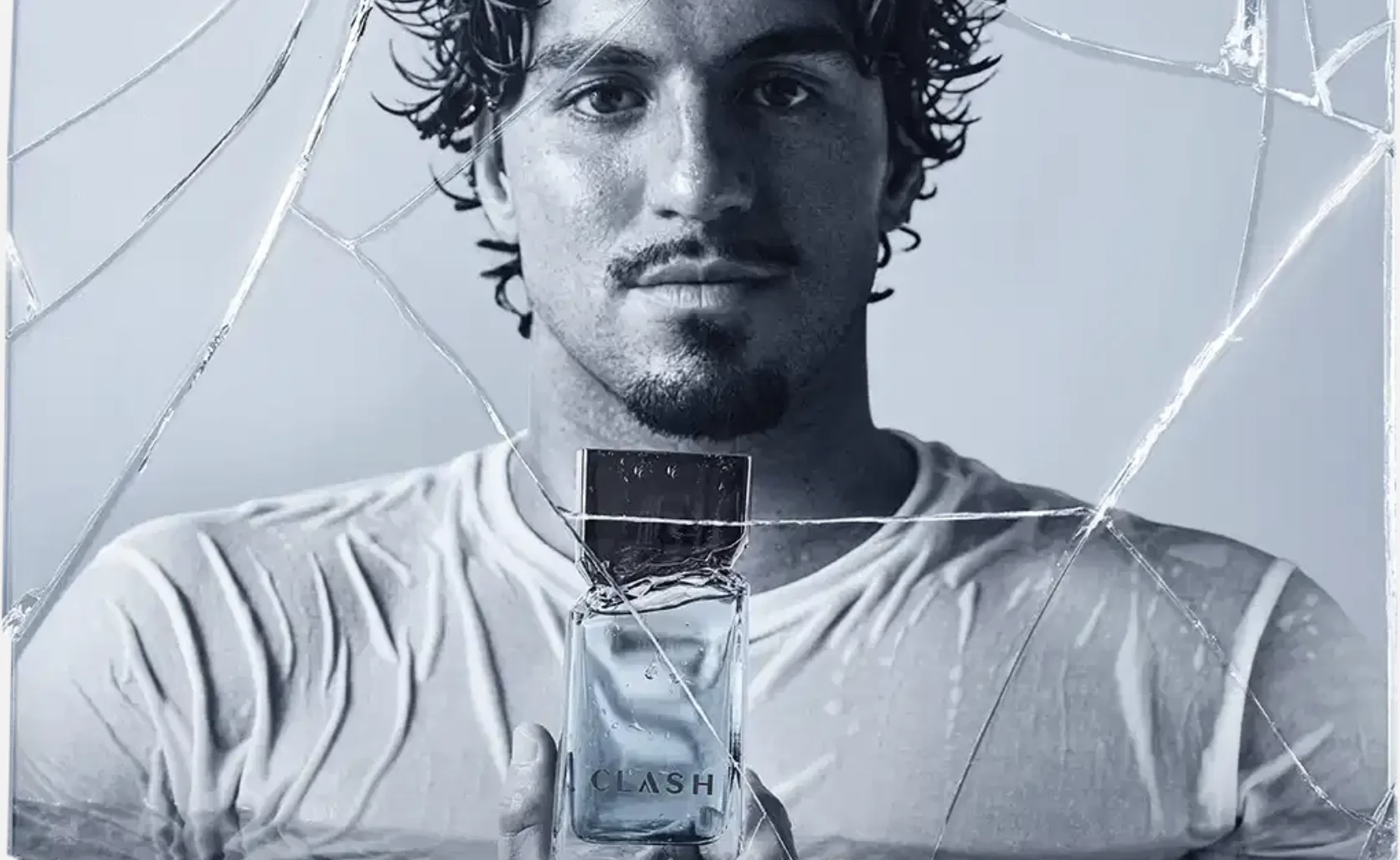 Homem de cabelos cacheados segura um frasco do perfume Clash diante de um efeito visual de vidro rachado.