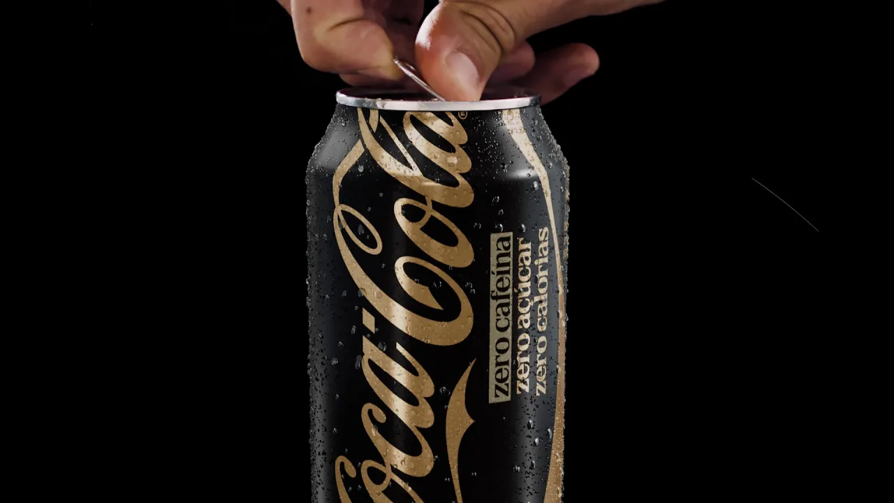Mão abrindo uma lata gelada de Coca-Cola Zero Cafeína, com embalagem preta e dourada sobre fundo escuro.